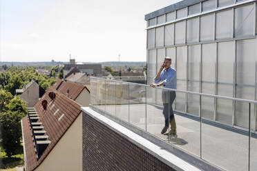 Geschäftsmann im Gespräch über Smartphone, während er auf dem Balkon gegen den Himmel an einem sonnigen Tag steht - JOSEF01807