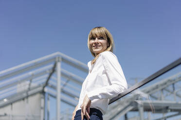 Lächelnde Unternehmerin, die an einem Geländer vor blauem Himmel steht und wegschaut - JOSEF01830