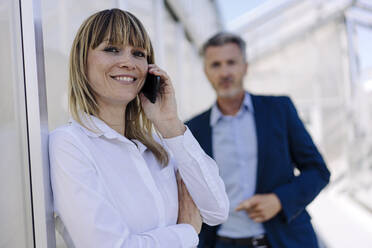 Smiling businesswoman talking over smart phone while male coworker standing in background - JOSEF01835