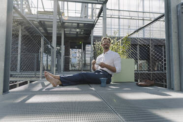 Smiling businessman with coffee sitting on floor in plant nursery - JOSEF01881