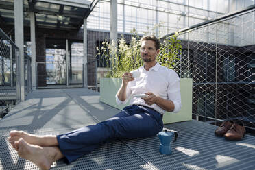 Male professional with coffee sitting on floor in plant nursery - JOSEF01882
