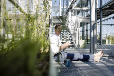 Male professional with coffee relaxing on floor in plant nursery - JOSEF01883