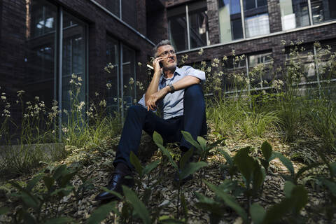 Businessman talking over smart phone while sitting amidst plants against building stock photo