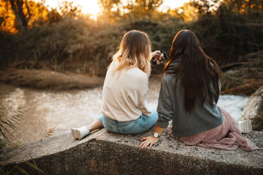 Sisters taking selfie while sitting on retaining wall in forest during sunset - MRRF00378