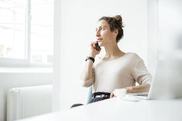 Geschäftsfrau, die über ein Smartphone spricht, während sie mit einem Laptop auf dem Tisch sitzt - JOSEF01893