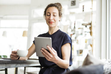 Businesswoman holding coffee cup using digital tablet while sitting at table - JOSEF01896