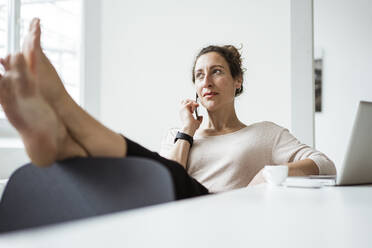 Geschäftsfrau mit Füßen auf dem Tisch, die beim Entspannen über ein Smartphone spricht - JOSEF01897