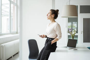 Thoughtful businesswoman holding smart phone standing by table - JOSEF01901