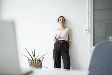 Confident businesswoman with hand in pocket holding coffee cup while standing against white wall - JOSEF01910