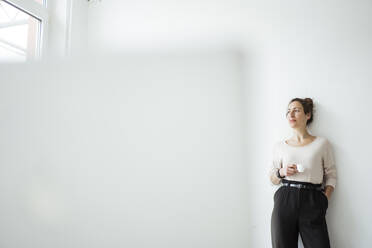 Businesswoman holding coffee cup contemplating while standing against white wall - JOSEF01911
