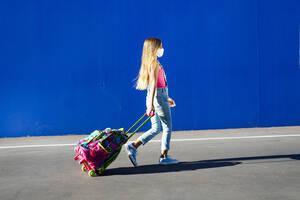 Blond girl walking with schoolbag on street against blue wall - JCMF01378