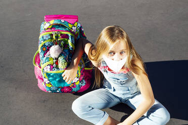 Mädchen sitzt auf einem Fußweg und trägt eine Schutzmaske an einem sonnigen Tag - JCMF01379