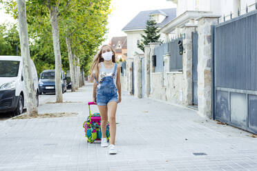 Girl with schoolbag walking on footpath wearing protective face mask - JCMF01380