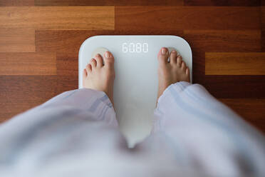 Top view crop faceless barefoot female in cozy pajama standing on digital weight and body fat scales with display showing healthy weight of 60 kg on bathroom floor in morning - ADSF15399