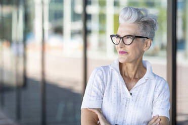 Smiling senior woman with arms crossed looking away while standing against glass window - WPEF03310