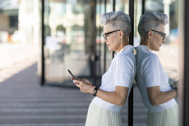 Reflektierendes Bild einer älteren Frau, die sich an ein Glasfenster lehnt, während sie ein Smartphone in der Stadt benutzt - WPEF03317