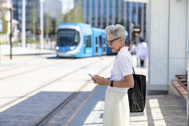 Ältere Frau schreibt eine SMS auf ihrem Smartphone, während sie in der Stadt an einer Straßenbahnhaltestelle steht - WPEF03325