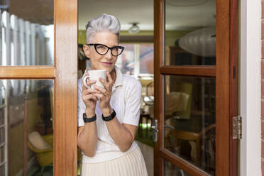 Ältere Frau lehnt an der Tür und hält eine Kaffeetasse in der Hand - WPEF03332