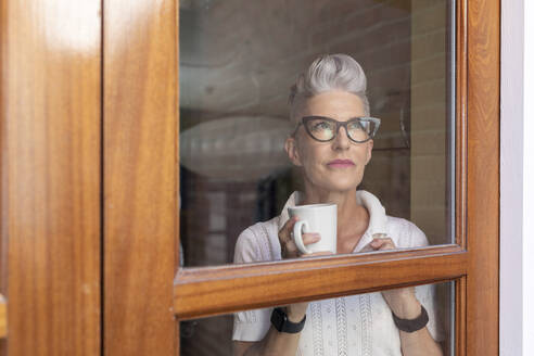 Ältere Frau schaut durch das Fenster, während sie zu Hause steht - WPEF03333