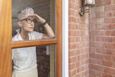 Ältere Frau schaut aus dem Fenster, während sie zu Hause steht - WPEF03337