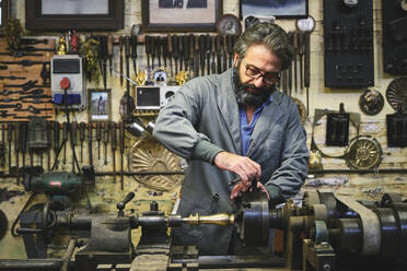 Fokussierter reifer bärtiger Goldschmied mit Brille bei der Arbeit an einer Poliermaschine in einer Werkstatt, während er einen spitzen Metallstab hält - ADSF15426