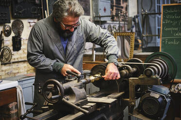 Ernster, reifer Juwelier in Uniform und mit Brille, der einen Metallstab an einer professionellen Ausrüstung während der Arbeit im Atelier schärft - ADSF15427