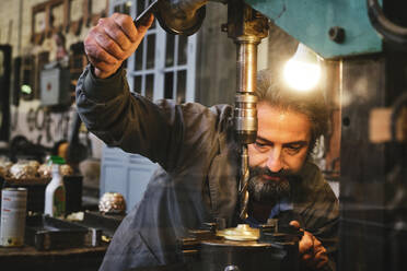 Konzentrierter Handwerker mittleren Alters in Uniform, der eine Graviermaschine benutzt, während er im Atelier mit Metall arbeitet - ADSF15428