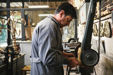 Seitenansicht eines reifen männlichen Juweliers in Arbeitskleidung beim Schleifen von Metallstäben auf einer Poliermaschine während der Arbeit in einer Werkstatt - ADSF15436