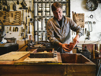 Ein erwachsener Handwerker in Arbeitskleidung füllt einen Formkasten mit Sand und bereitet sich auf den Metallguss in einer Goldschmiedewerkstatt vor - ADSF15441
