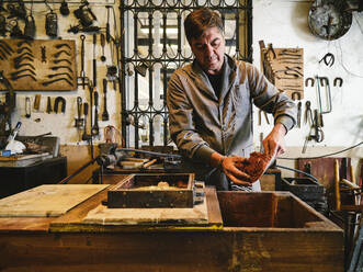 Skilled adult male craftsman in workwear filling molding box with sand while preparing for metal casting process in goldsmith workshop - ADSF15441