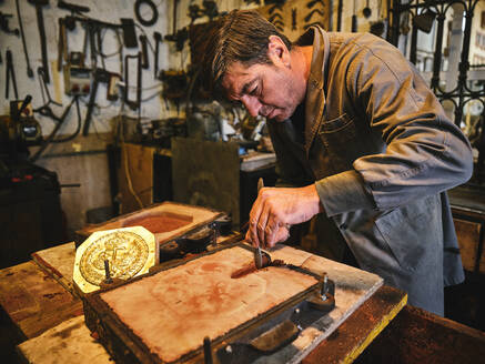 Konzentrierter reifer Handwerker beim Schneiden von Sandformen während der Arbeit mit Metallguss in der Goldschmiedewerkstatt - ADSF15443