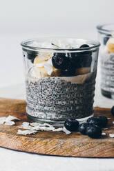 Glasgefäße mit köstlichem Chia-Pudding aus frischer leckerer Mandelmilch und Chia-Samen mit Honig, gekrönt mit Brombeeren und geschnittener Banane, serviert auf einem Holzbrett vor weißem Hintergrund - ADSF15451