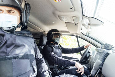Policemen in medical mask and protective gear talking on radio set while sitting in car with partner - ADSF15456