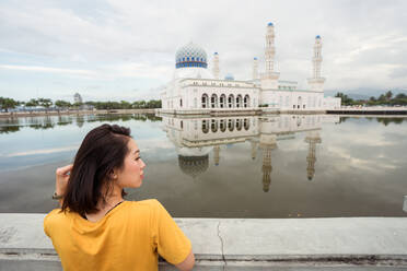 Back view of ethnic female tourist leaning on border near lake in front of famous Bandaraya Mosque and looking away during summer vacation in Kota Kinabalu - ADSF15462