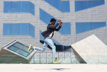 Full body side view of confident young African American businessman in trendy outfit running fast and jumping against contemporary city building - ADSF15505