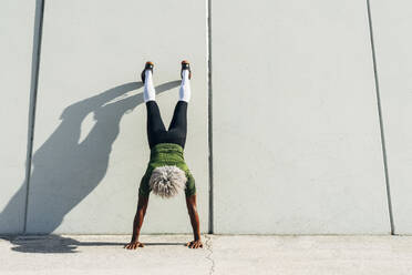 Back view of unrecognizable black sportsman with blond hair doing handstand near wall of modern building in city - ADSF15512