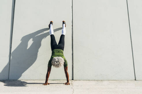 Back view of unrecognizable black sportsman with blond hair doing handstand near wall of modern building in city - ADSF15512