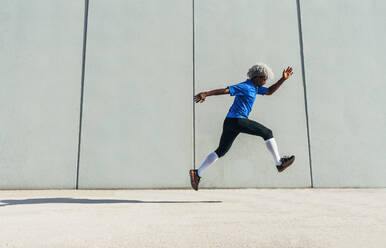 Ganzkörper-Seitenansicht eines schwarzen Sportlers in Sportkleidung, der während eines Fitness-Trainings an einem sonnigen Tag auf einer Straße in der Nähe eines modernen Gebäudes springt - ADSF15520