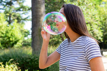 Seitenansicht einer sorglosen Frau, die im grünen Garten steht und Seifenblasen bläst, während sie das Sommerwochenende genießt - ADSF15529