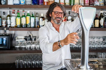 Erwachsener ethnischer Mann in weißem Hemd gießt Alkohol aus einer Biersäule in ein Glas, während er in einer Bar steht - ADSF15542