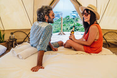 Full length happy mature couple in casual summer wear sitting on bed in luxury camping resort and looking at each other with tenderness - ADSF15544