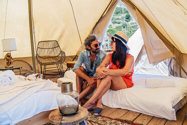 Full length happy mature couple in casual summer wear sitting on bed in luxury camping resort and looking at each other with tenderness - ADSF15545