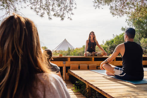 Rückenansicht einer nicht erkennbaren Person, die auf einer hölzernen Plattform die Yogastellung Padmasana einnimmt, während eines Trainings im Freien mit einer Lehrerin im Garten - ADSF15547