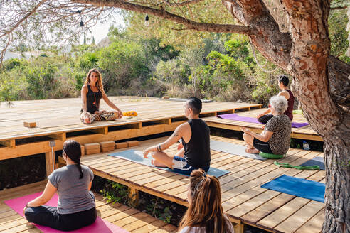 Rückenansicht einer nicht erkennbaren Person, die auf einer hölzernen Plattform die Yogastellung Padmasana einnimmt, während eines Trainings im Freien mit einer Lehrerin im Garten - ADSF15548