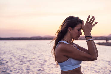 Side view of tranquil young female in sports bra meditating with hands near forehead against calm river and sunset sky in summer evening in nature - ADSF15559
