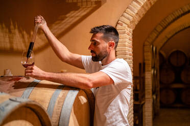 Side view serious male winemaker pouring aromatic red wine from wooden barrel into glass by using special glass tool - ADSF15567