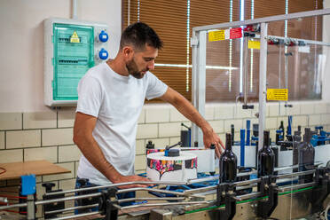 Seitenansicht professioneller Winzer mit weißem Hemd, der in einer modernen Fabrik arbeitet und Flaschen von einem automatischen Förderband einsammelt - ADSF15569