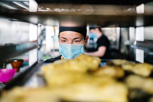 Concentrated female cook in hat and respirator standing against steel pan rack loaded with food while working in modern restaurant kitchen - ADSF15574