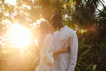Loving multiracial couple in white shirts bonding and kissing tenderly while enjoying evening in summer garden in sunset time in Madrid Spain - ADSF15606