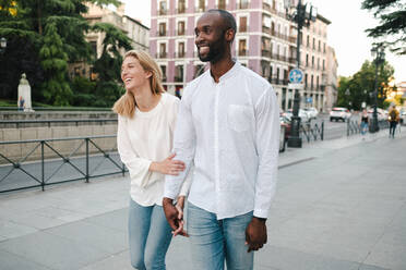 Optimistisches lächelndes gemischtrassiges Paar in Jeans und weißen Hemden, das eine moderne Straße in Madrid entlanggeht und sich zärtlich an den Händen hält - ADSF15608